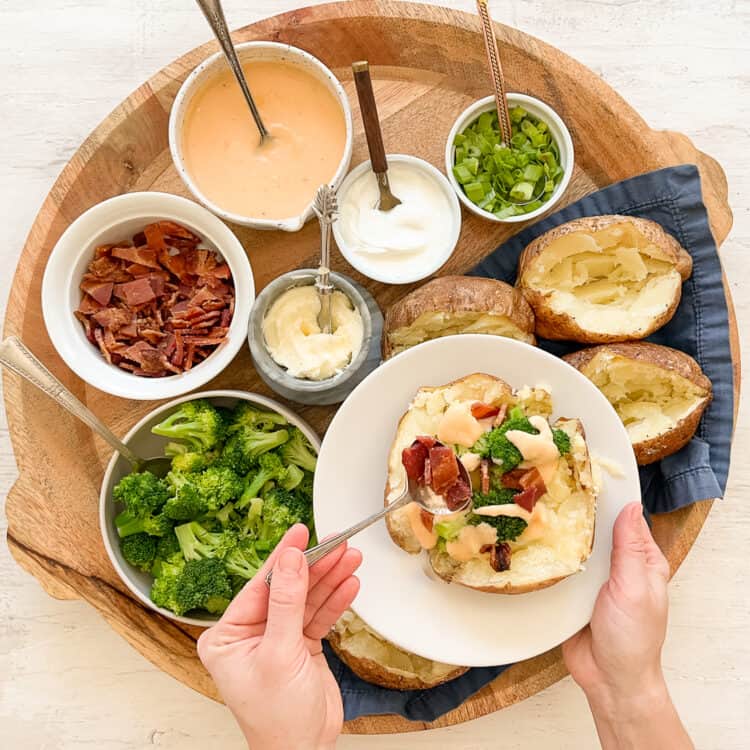 Someone holding a plate with a baked potato topped with homemade cheese sauce and steamed broccoli and using a spoon to add chopped bacon to the top.