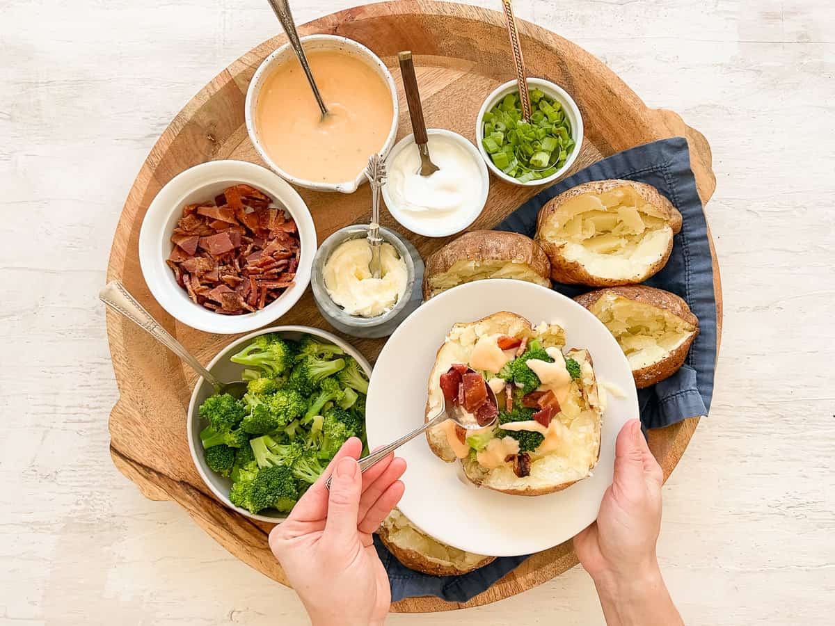 Someone holding a plate with a baked potato topped with homemade cheese sauce and steamed broccoli and using a spoon to add chopped bacon to the top.