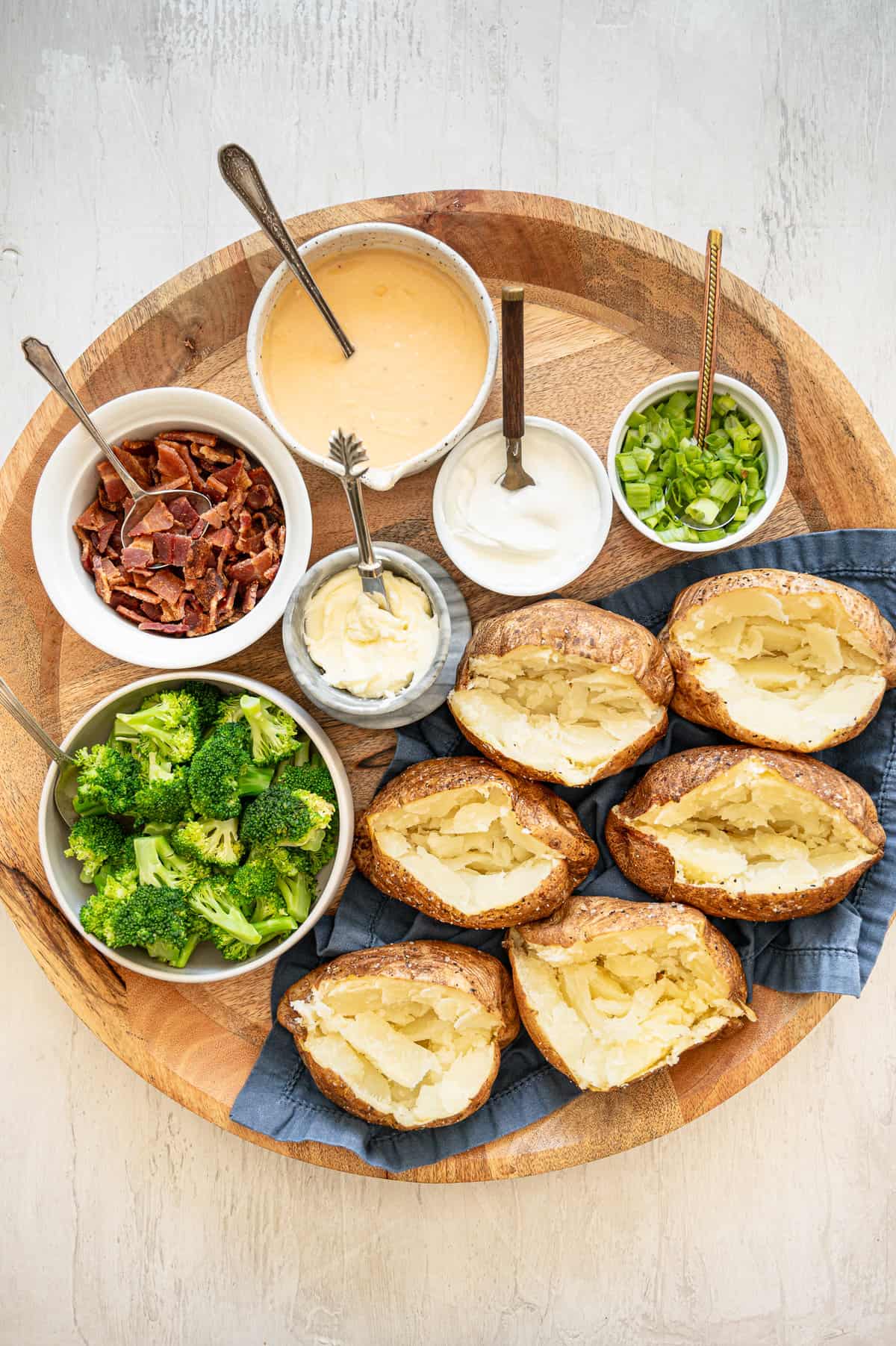 Baked potatoes and multiple toppings including chopped broccoli, homemade cheese sauce, sour cream, butter, chopped bacon, and scallions all laid out on a circular wooden cutting board.