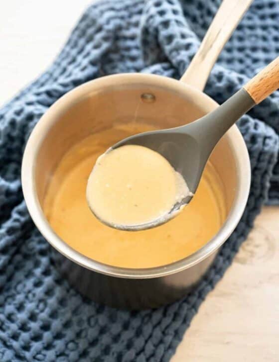 Homemade cheese sauce in a small saucepan with some being lifted out with a spoon.