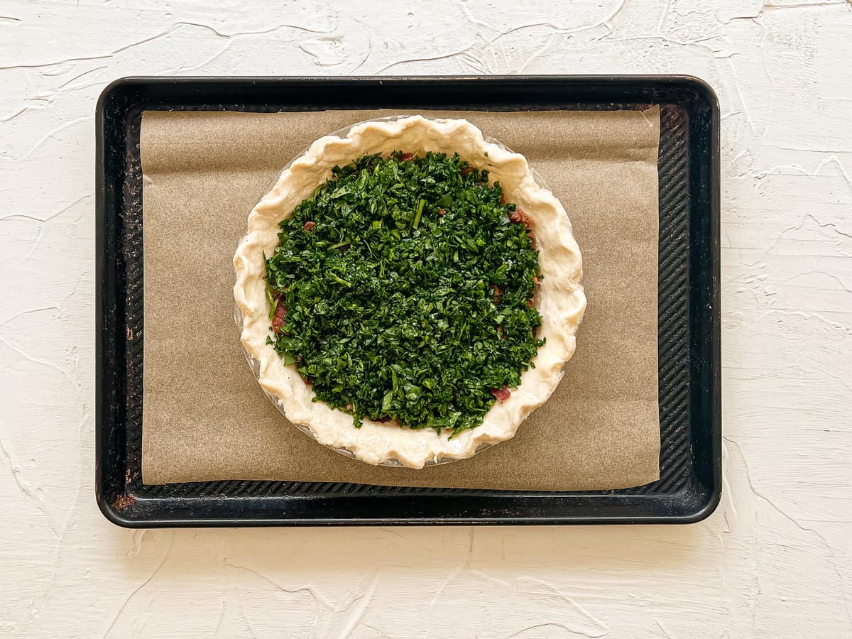An uncooked pie crust with chopped spinach on top of a layer pf chopped bacon sitting on a baking sheet with parchment paper.