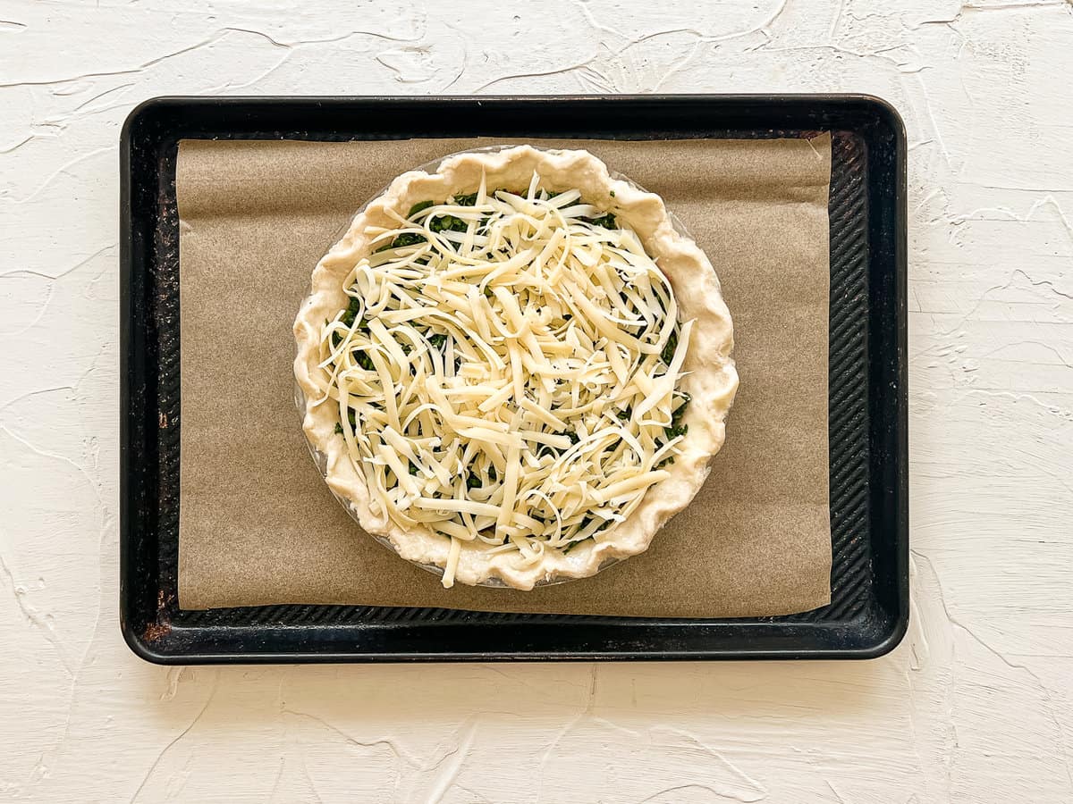 An uncooked pie crust on a baking sheet with parchment paper filled with layers of chopped bacon, chopped spinach, and shredded swiss cheese.