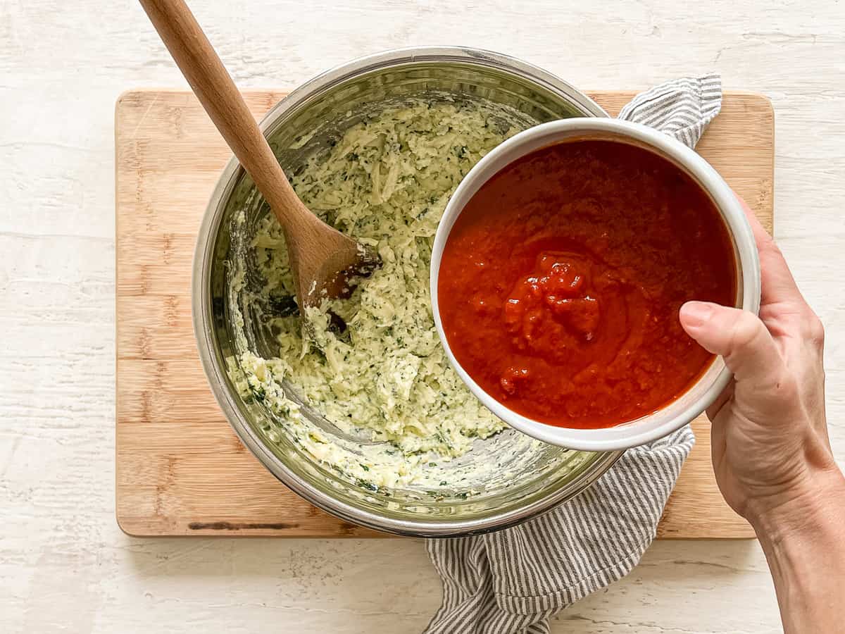 A mixing bowl with three cheeses, eggs, and seasonings mixed together and someone holding a bowl of marinara sauce to pour in.