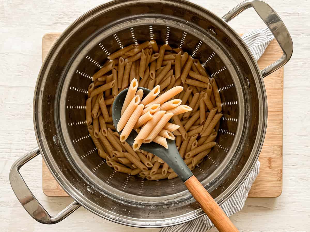A wooden spoon lifting cooked penne pasta out of a metal colander.