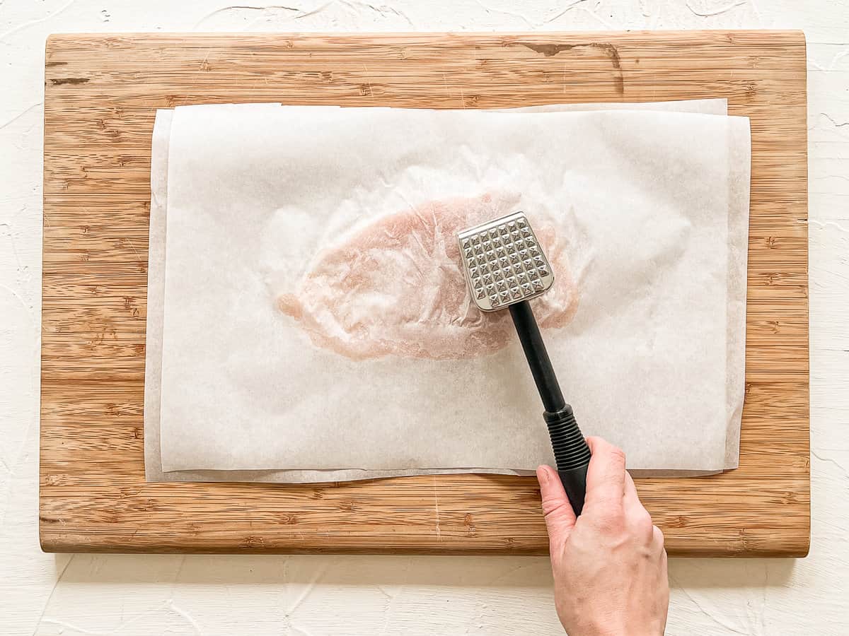 Chicken breast between two pieces of parchment paper on a wooden cutting board with a hand using a kitchen mallet to make it thinner.