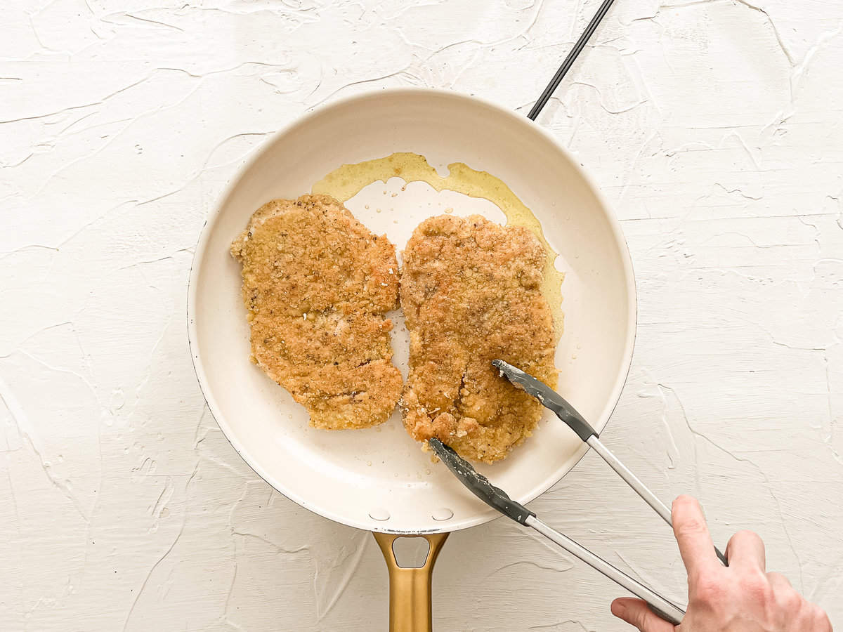 Two breaded chicken cutlets frying in oil in a ceramic frying pan.