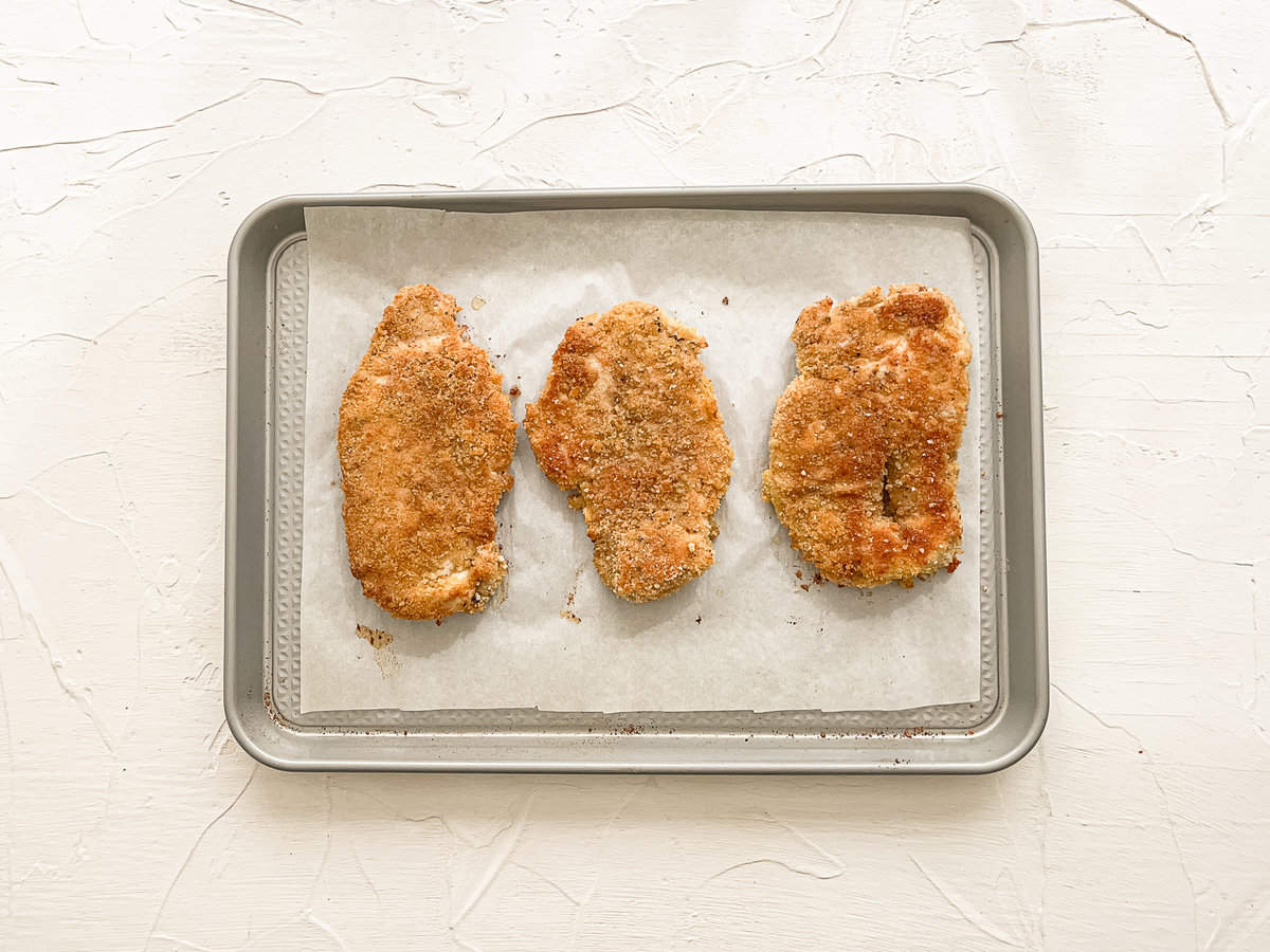Three chicken cutlets sitting on parchment paper on a baking sheet.