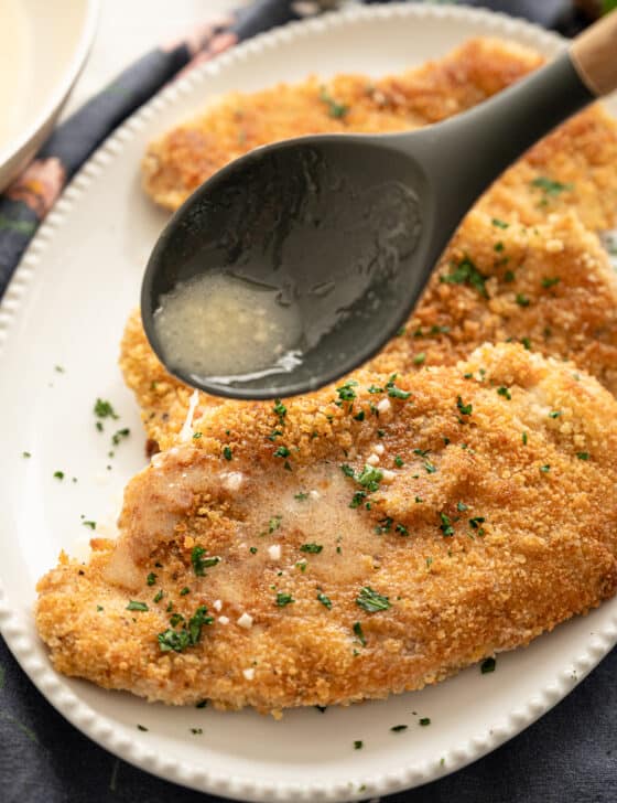 Spoon drizzling lemon butter sauce over crispy chicken cutlets on a white serving platter.
