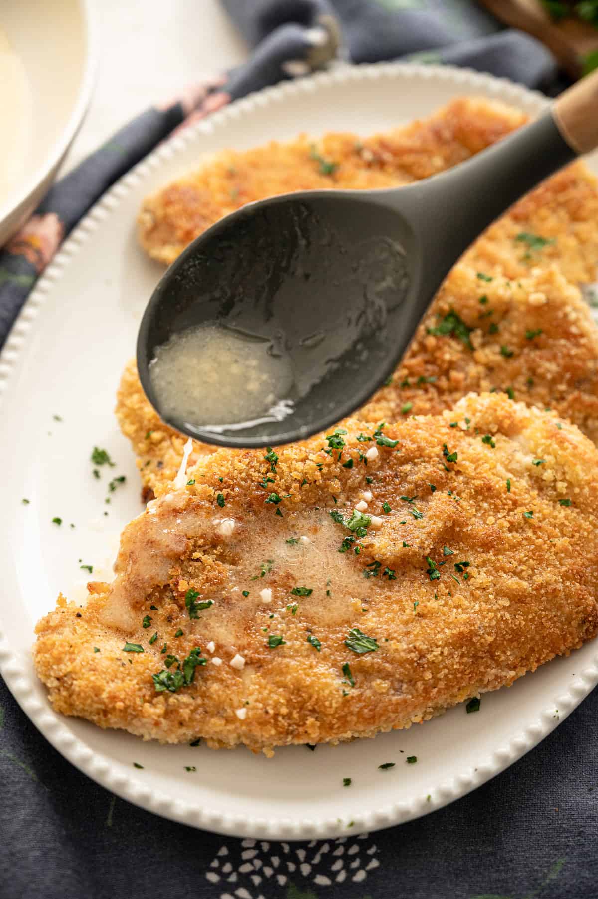 Spoon drizzling lemon butter sauce over crispy chicken cutlets on a white serving platter.