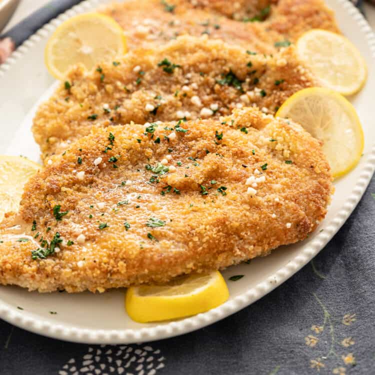 Crispy chicken cutlets on a serving platter with lemon butter sauce over top and lemon slices around the outside of the platter.