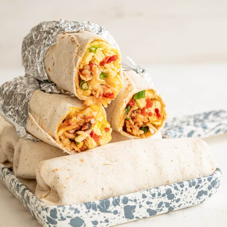 Breakfast burritos lined up in a marbled serving dish with three on top that have the ends cut off to see the scrambled eggs, bacon, peppers, and green onions.