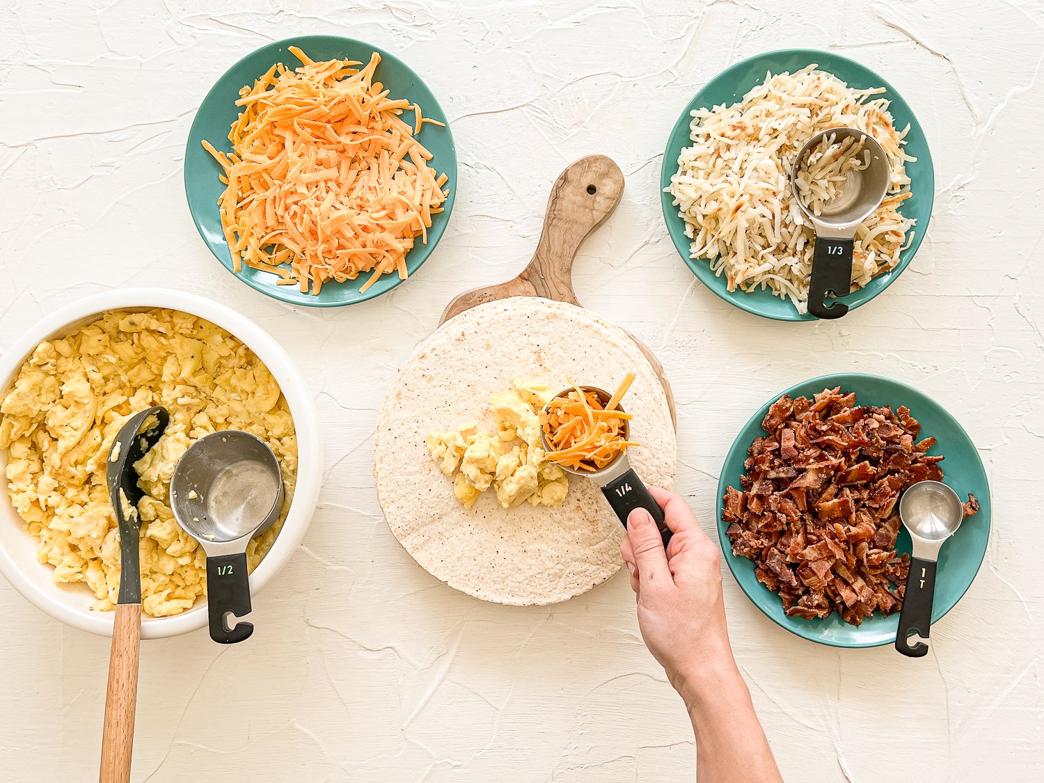 Breakfast burrito prep station with scrambled eggs, shredded cheddar cheese, shredded hash browns, chopped bacon, and tortillas with a hand putting a scoop of cheddar cheese on a tortilla.