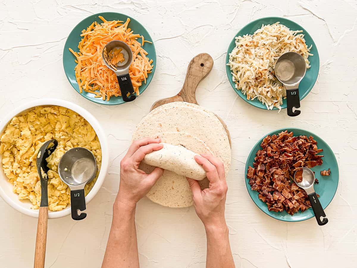 Breakfast burrito prep station with bowls of ingredients in a semi-circle and a hand rolling up a filled tortilla.