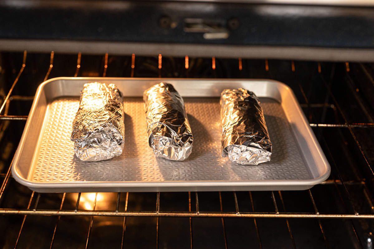Three breakfast burritos wrapped in foil on a baking sheet in the oven.