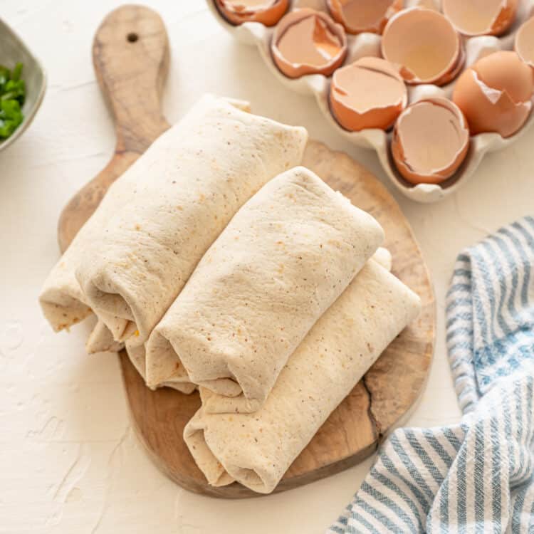 A stack of breakfast burritos on a wooden cutting board with eggshells off to the side.