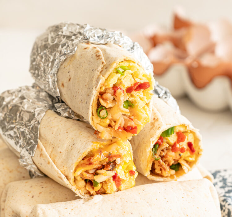 Stack of breakfast burritos with the top ones having the ends cut off so you can see the scrambled eggs, bacon, sauteed peppers, and more inside.