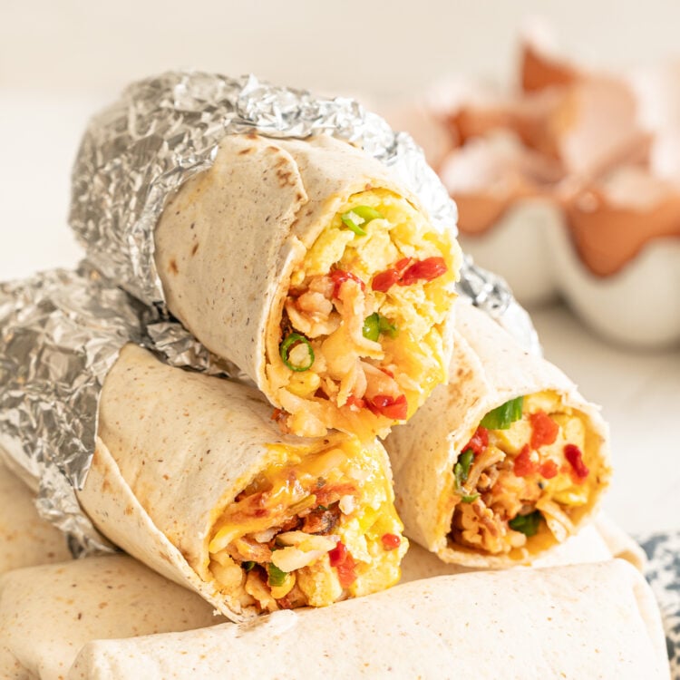 Stack of breakfast burritos with the top ones having the ends cut off so you can see the scrambled eggs, bacon, sauteed peppers, and more inside.
