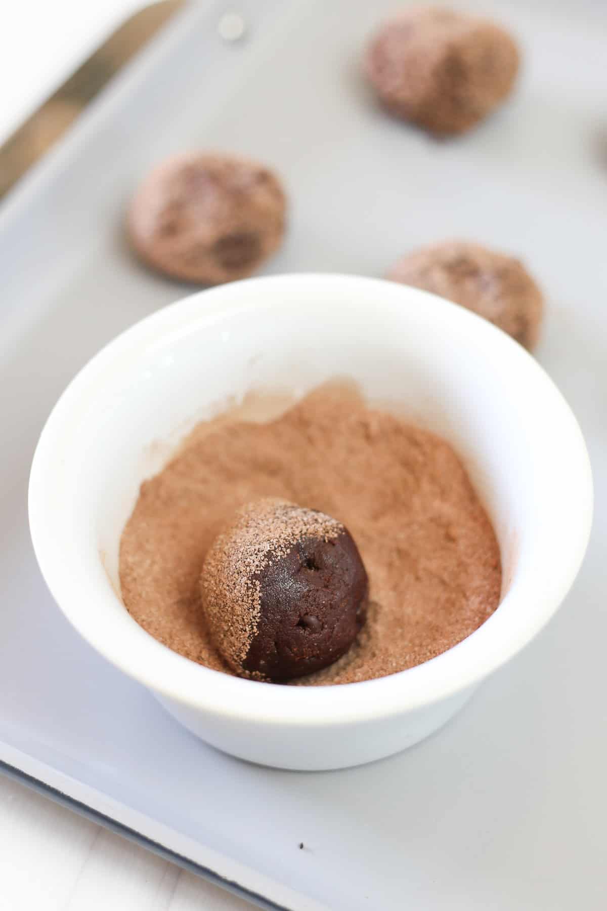 A small white bowl of cocoa and sugar mixed together with a brownie mix cookie dough ball being rolled in it.