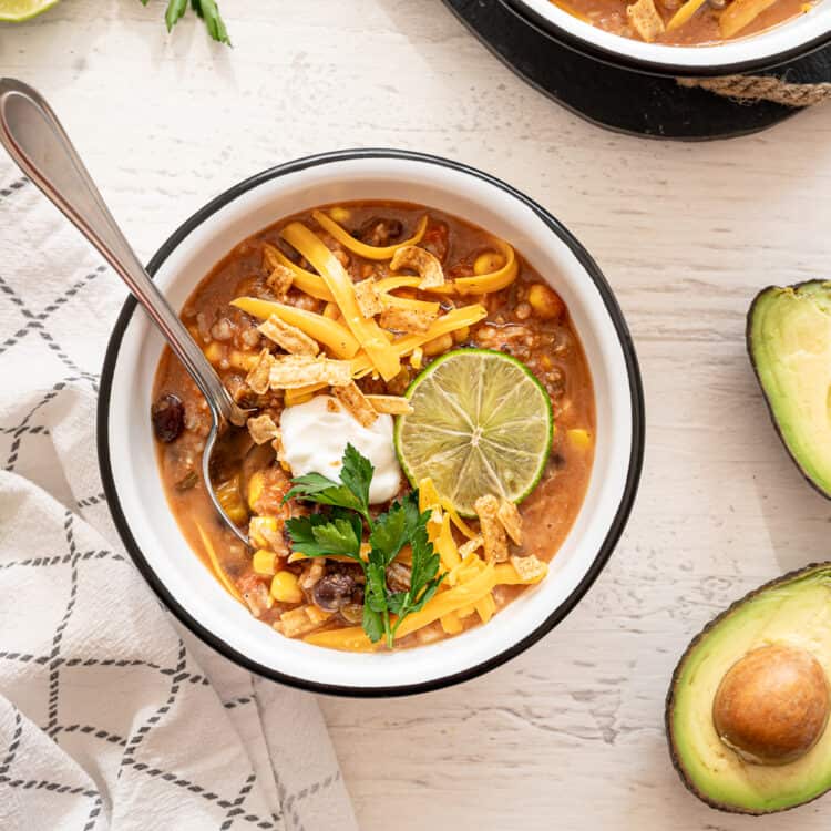 Bowl of vegetarian tortilla soup with lots of toppings on it as well as lime wedges and an avocado cut in half sitting beside it.