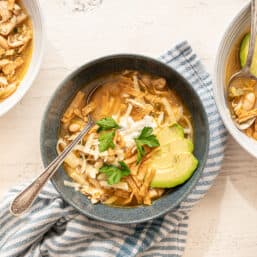 Three bowls of white chicken chili with avocado slices, sour cream, tortilla strips, fresh cilantro, and shredded cheese on top.