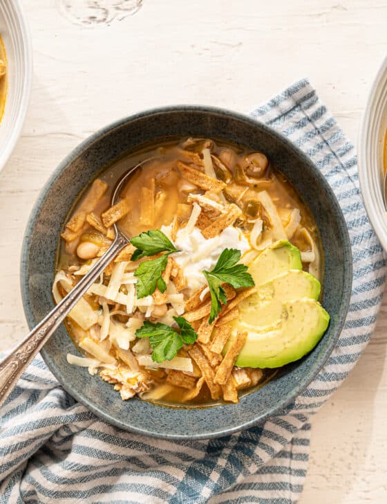 Three bowls of white chicken chili with avocado slices, sour cream, tortilla strips, fresh cilantro, and shredded cheese on top.