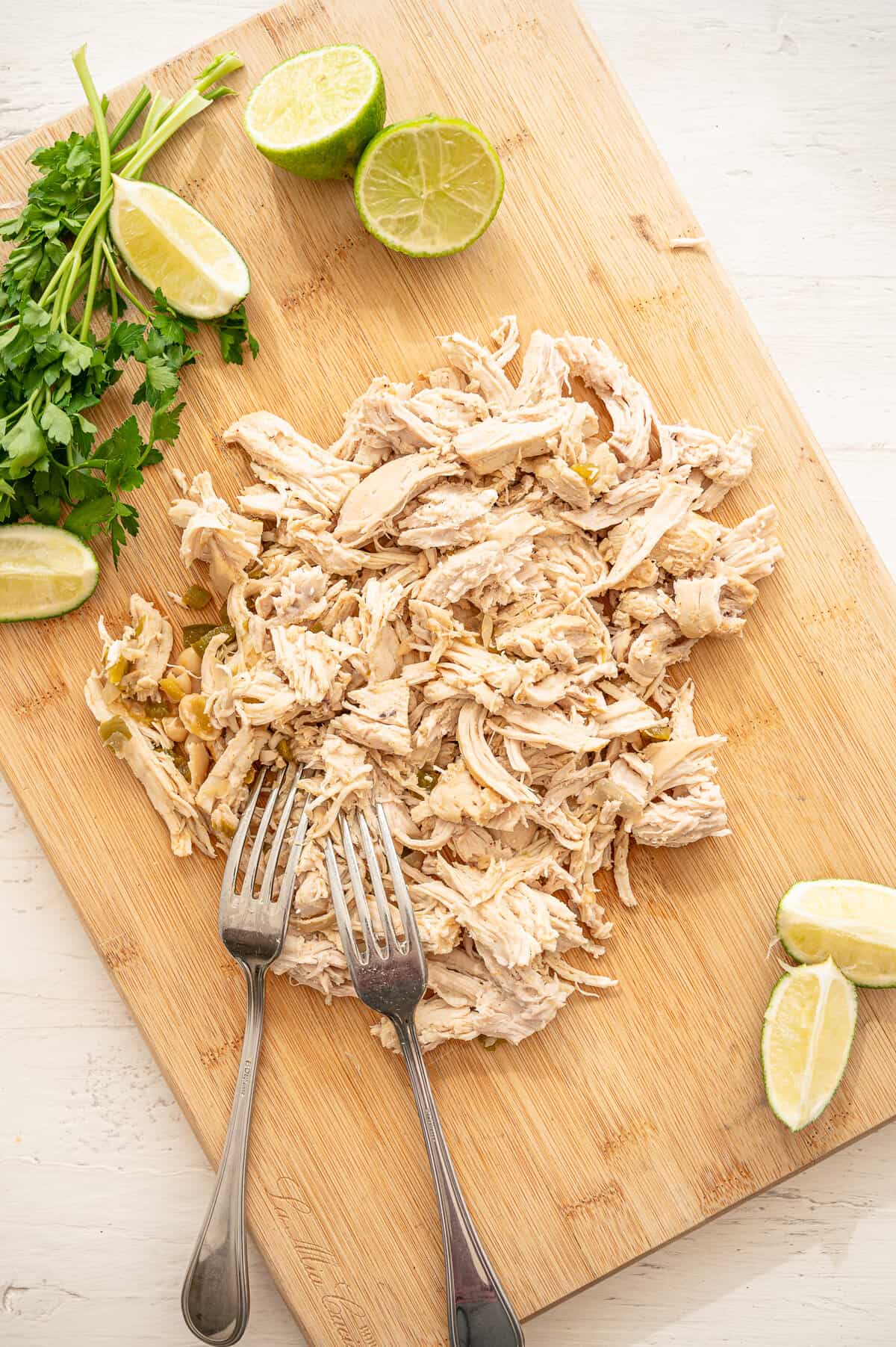 Shredded chicken on a wooden cutting board with two forks and lime wedges as well.