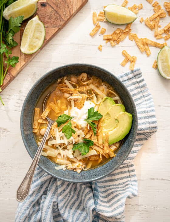 A bowl of white chicken chili with tortilla strips, shredded cheese, a dollop of sour cream, avocado slices, and fresh cilantro on top.