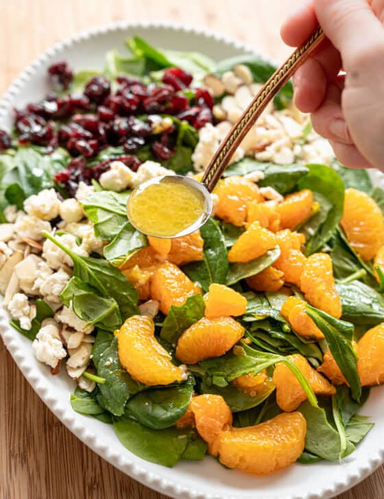 Creamy citrus dressing being ladled onto Mandarin Orange Spinach Salad with sliced almonds, dried cranberries, and feta cheese on it.