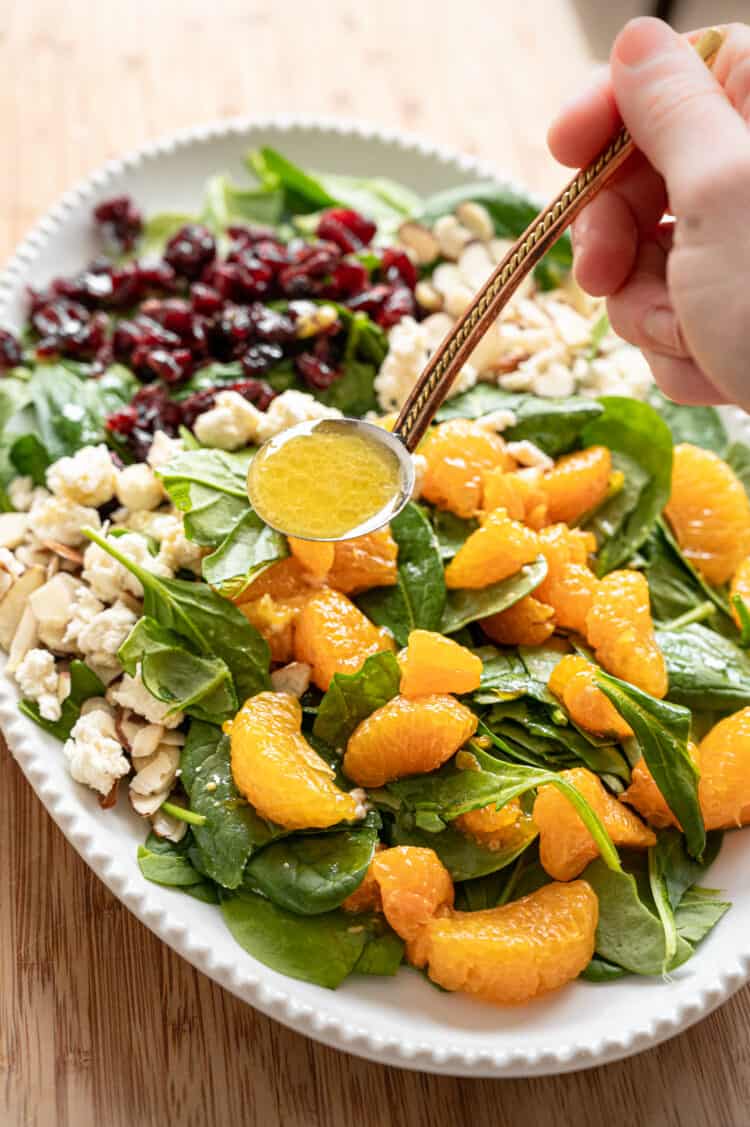 Creamy citrus dressing being ladled onto Mandarin Orange Spinach Salad with sliced almonds, dried cranberries, and feta cheese on it.