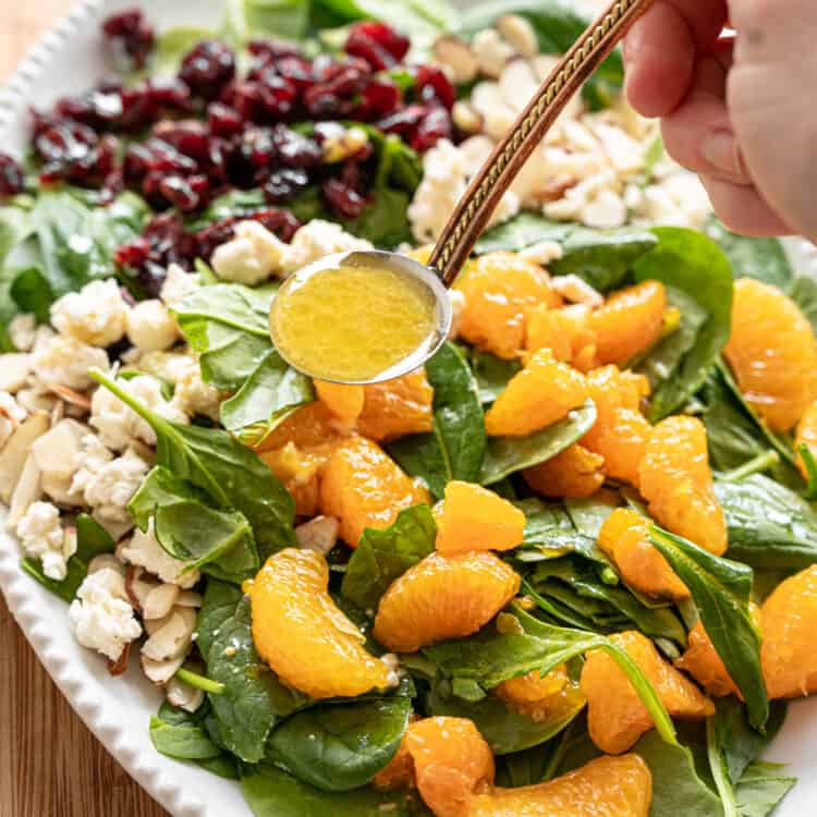 Creamy citrus dressing being ladled onto Mandarin Orange Spinach Salad with sliced almonds, dried cranberries, and feta cheese on it.