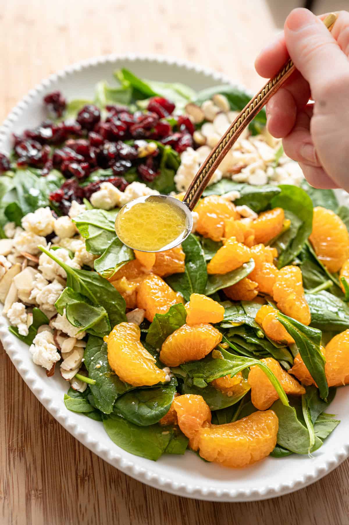 Creamy citrus dressing being ladled onto Mandarin Orange Spinach Salad with sliced almonds, dried cranberries, and feta cheese on it.