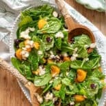 Mandarin orange spinach salad ingredients including sliced almonds, feta cheese, and dried cranberries tossed in a creamy citrus dressing on a white platter.