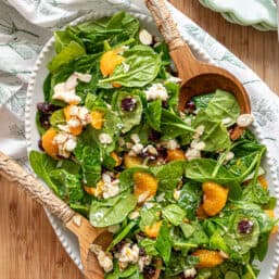 Mandarin orange spinach salad ingredients including sliced almonds, feta cheese, and dried cranberries tossed in a creamy citrus dressing on a white platter.