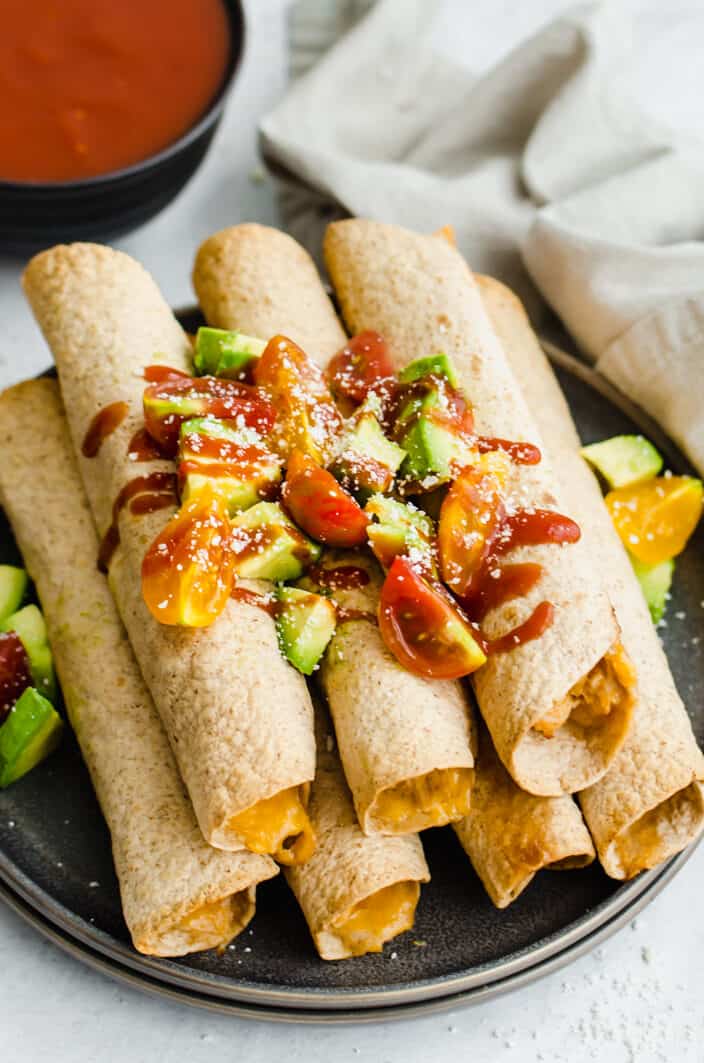 Chicken taquitos stacked up on a plate with chopped avocado and tomatoes on top.