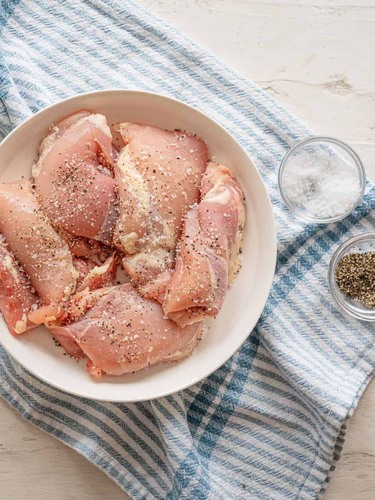 Seasoned raw chicken thighs on a plate with small bowls of salt and pepper next to it.