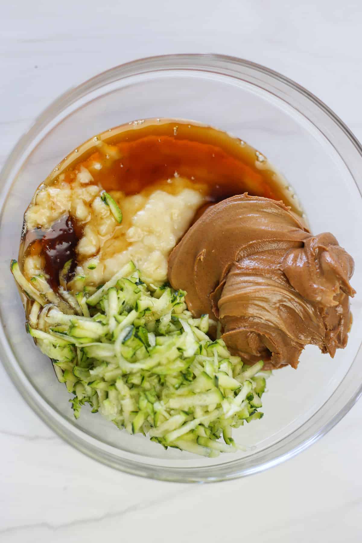 Mashed banana, shredded zucchini, real maple syrup, and creamy peanut butter in a glass bowl ready to be mixed.