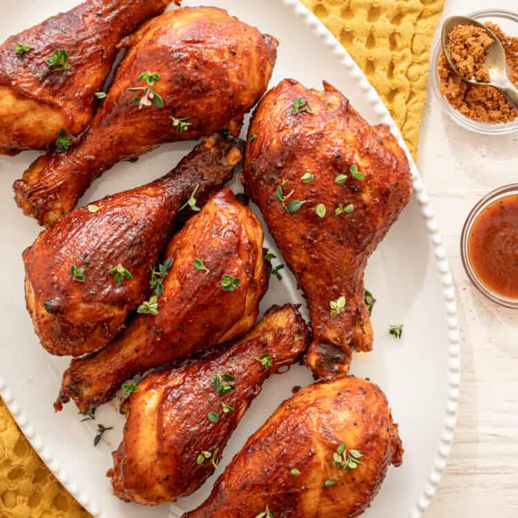 BBQ chicken drumsticks lined up on a white platter with fresh thyme sprinkled on top.