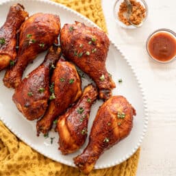 BBQ chicken drumsticks lined up on a white platter with small bowls of BBQ sauce and dry rub beside it.