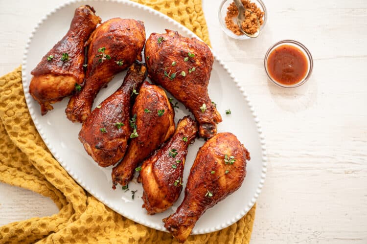 BBQ chicken drumsticks lined up on a white platter with small bowls of BBQ sauce and dry rub beside it.