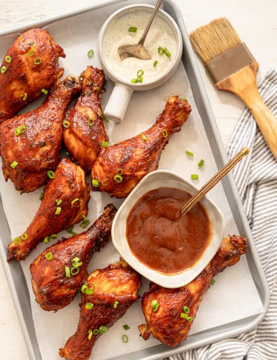 BBQ chicken drumsticks lined up on a parchment paper lined baking sheet with bowls of ranch and BBQ sauce.
