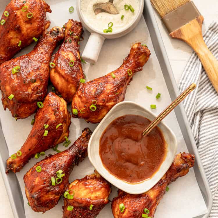 BBQ chicken drumsticks lined up on a parchment paper lined baking sheet with bowls of ranch and BBQ sauce.