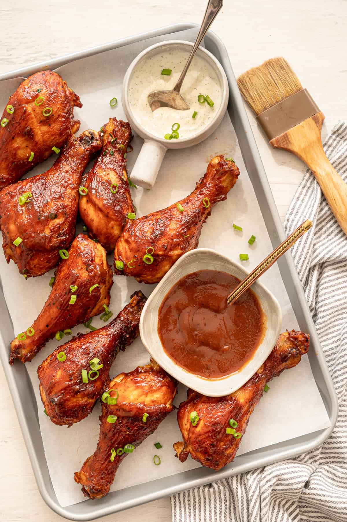 BBQ chicken drumsticks lined up on a parchment paper lined baking sheet with bowls of ranch and BBQ sauce.