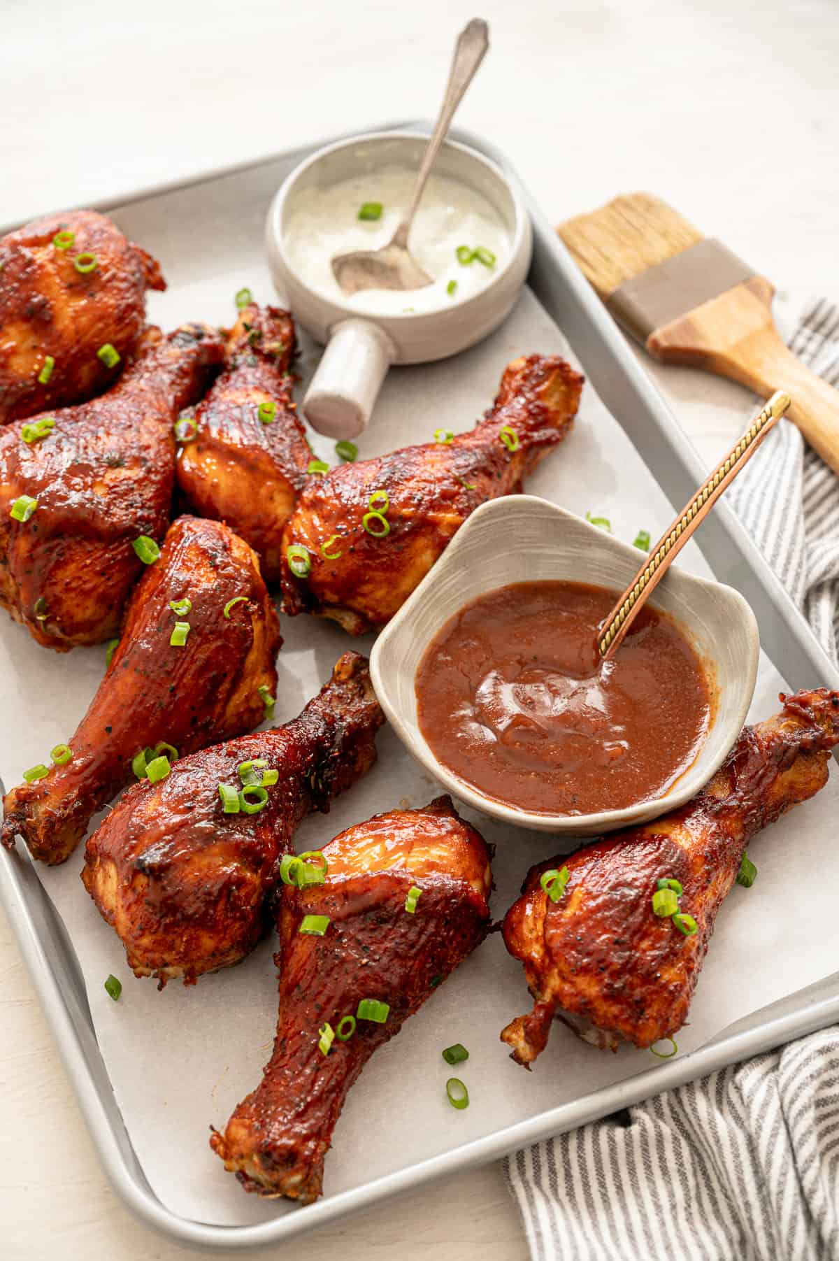 BBQ chicken drumsticks sprinkled with fresh chives lined up on a baking sheet with bowls of ranch and BBQ sauce.