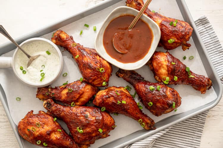 BBQ chicken drumsticks lined up with bowls of ranch and BBQ sauce on the baking sheet too.