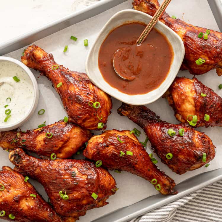 BBQ chicken drumsticks lined up with bowls of ranch and BBQ sauce on the baking sheet too.