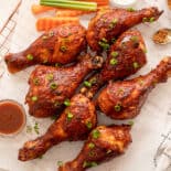 BBQ chicken drumsticks lined on parchment paper served with extra BBQ sauce and crinkle cut celery and carrots.