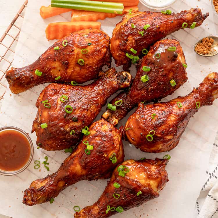 BBQ chicken drumsticks lined on parchment paper served with extra BBQ sauce and crinkle cut celery and carrots.
