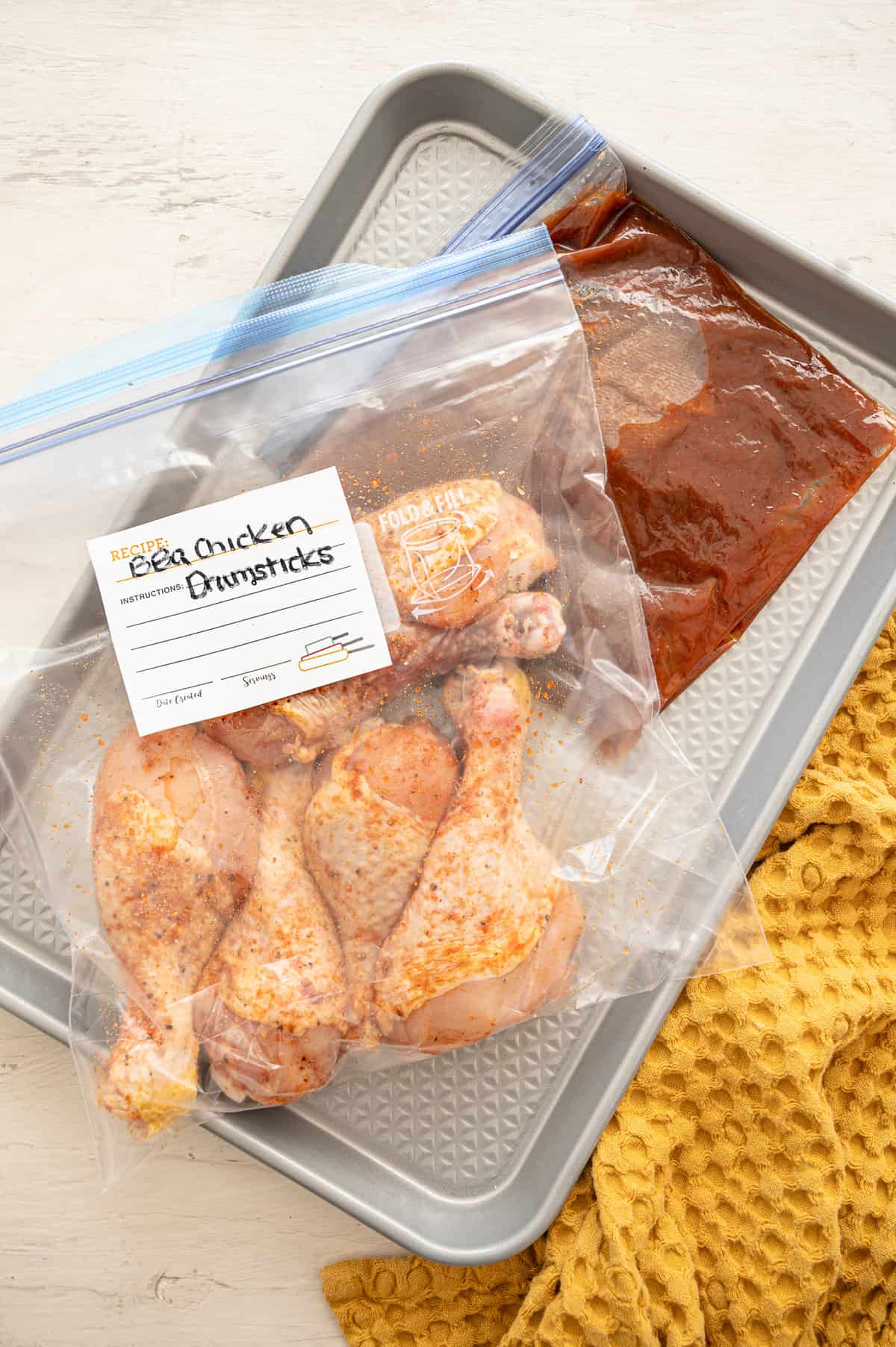 Chicken drumsticks with dry rub in a gallon-size freezer bag with a smaller bag of BBQ sauce laid on a baking sheet.