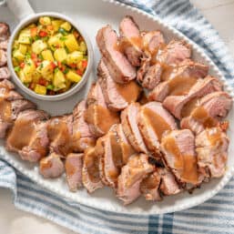 A white platter of sliced grilled pork tenderloin with Hawaiian marinade sauce drizzled over it.