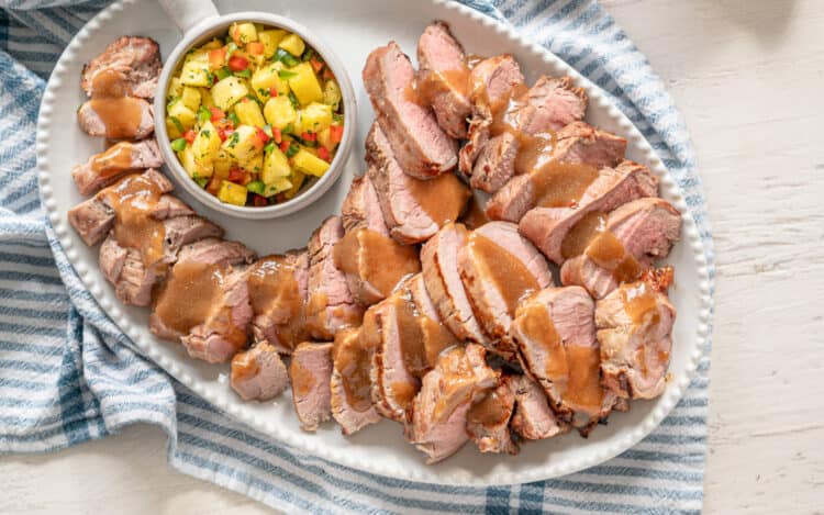 A white platter of sliced grilled pork tenderloin with Hawaiian marinade sauce drizzled over it.